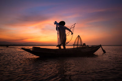 Silhouette fisherman throwing fishing net in sea while standing on boat during sunset
