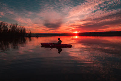 Silhouette person sitting in kayak on lake against dramatic sky during sunset