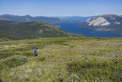 Rear view of person riding bicycle on field against river and mountains