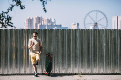 Young man with cell phone leaning against a wall next to longboard with city in background