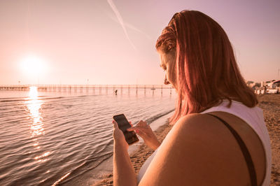 Mature woman using mobile phone at beach against sky during sunset
