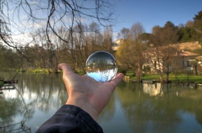 Cropped image of person hand holding crystal ball by river against sky