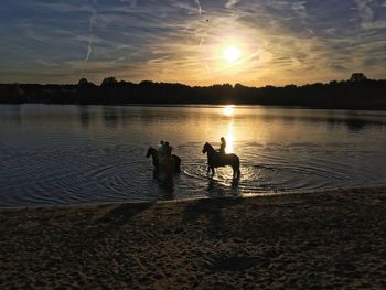 Silhouette people on beach by lake against sky during sunset