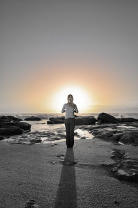 Full length rear view of woman standing on shore at beach against sky during sunset