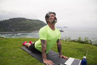 Mature man exercising on green grass with yoga mat outdoors at sunset