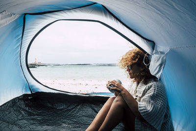 Young woman looking at camera while sitting by sea against sky