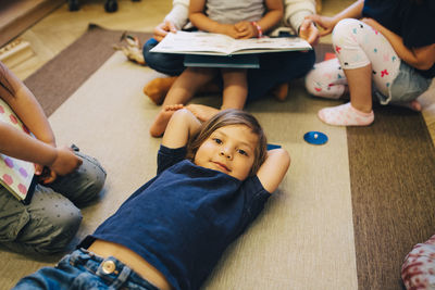 High angle portrait of boy lying on carpet amidst friends in classroom