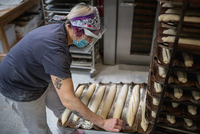 High angle side view of female worker in mask and shield making notches on loaves of baguette dough while preparing pastry for baking