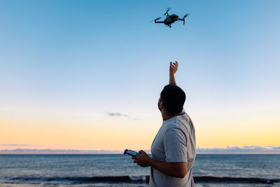 Unrecognizable black male in casual clothes launching modern drone against sunset sky while spending time on beach near waving sea