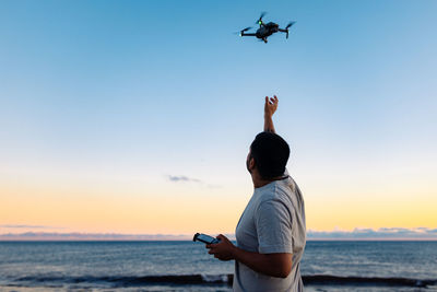 Rear view of man standing at beach against sky during sunset