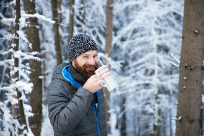 Portrait of mature man drinking coffee while standing against snow covered trees during winter