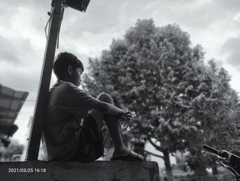 Side view of man sitting by tree against sky