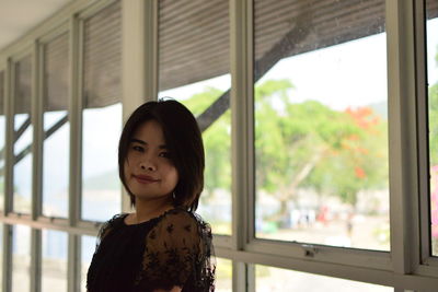 Portrait of young woman standing against window at home