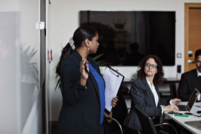 Businesswoman holding clipboard while discussing with colleagues during meeting in office