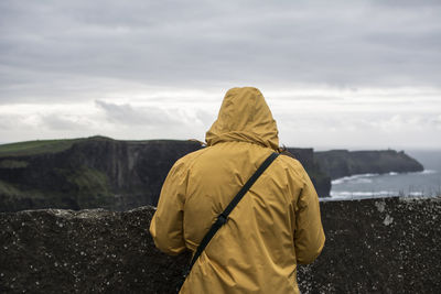 Rear view of woman wearing yellow warm clothing with cliff in background against cloudy sky