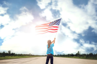 Rear view of girl holding american flag while standing on road against sky