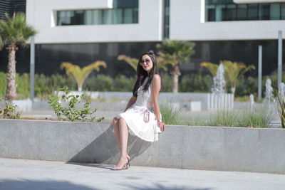 Full length portrait of woman wearing sunglasses sitting on retaining wall