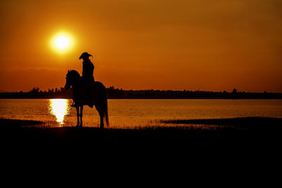 Silhouette people riding by lake against orange sky