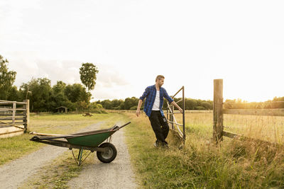 Man opening gate by wheelbarrow on dirt road against sky at farm