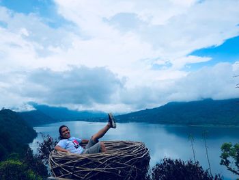 Portrait of man sitting in large nest with lake in background against cloudy sky