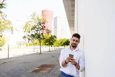 Young bearded man leaning against a white wall using the mobile phone