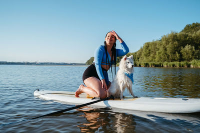 Happy young woman enjoying life on the lake at early morning sitting on the sup board with her dog
