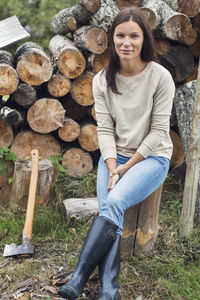 Full length portrait of woman sitting on tree stump against stacked firewood at yard