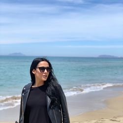 Young woman wearing sunglasses standing at beach against sky