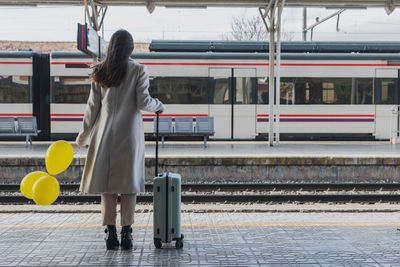 Back view of unrecognizable female traveler in stylish outfit standing with yellow balloons in hand and suitcase on platform of railway station