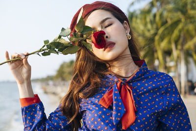 Close-up of beautiful young woman holding rose standing on beach against sky