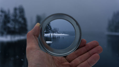 Cropped hand holding glass object against lake during winter