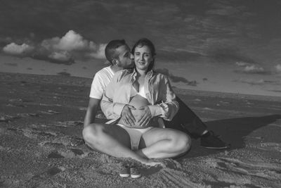 Man with pregnant woman sitting at beach against sky