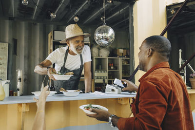 Smiling owner serving food while male customer doing contactless payment through smart phone