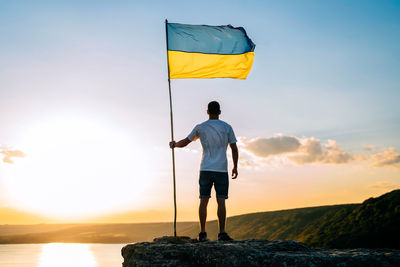 Rear view of woman holding flag against sky during sunset