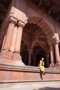 Full body happy asian lady in yellow dress looking at camera while sitting in large arch of aged red fort on sunny day in new delhi, india