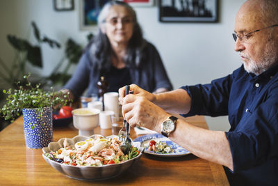 Senior man serving salad for himself while sitting with woman at table