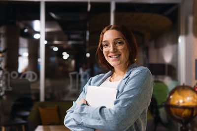 Cheerful young businesswoman in suit and glasses embracing netbook and looking at camera with smile in workspace