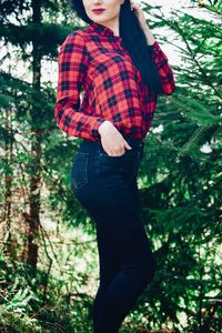 Midsection of young woman with hand in pocket standing by tree at forest