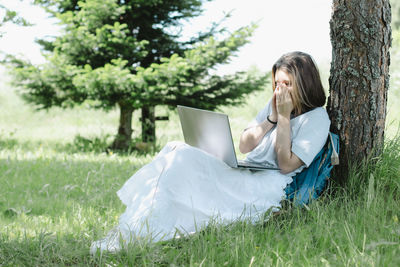 Nice teenage girl siting near tree in summer park with open laptop in her hands