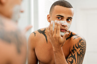 Shirtless young man applying moisturizer in front of mirror at home