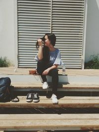 Full length of smiling young woman sitting against wall during sunny day