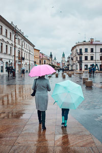 Back view of mother and her daughter holding the pink and blue umbrellas walking in a downtown