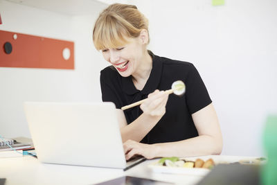 Smiling businesswoman eating lunch while using laptop in office