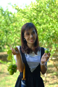 Portrait of smiling young woman holding limes while standing against trees in farm