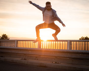 Man jumping on railing against sky during sunset