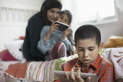 Boy using digital tablet in bedroom with family in background