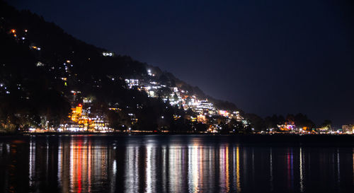 Illuminated buildings by lake against sky at night
