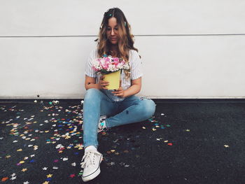 Full length of woman holding flower pot while sitting against wall