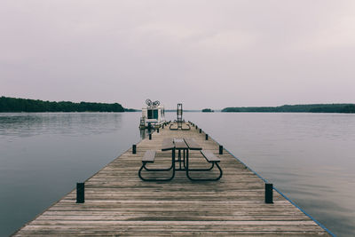 Bench and table on pier at lake against cloudy sky