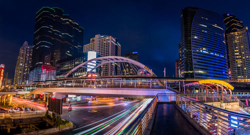 Light trails on bridge by buildings against sky at night